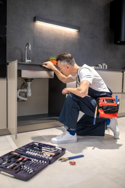 Household repair. Middle-aged man in blue overalls knelt down with hand touching sink carefully examining pipes in kitchen at home
